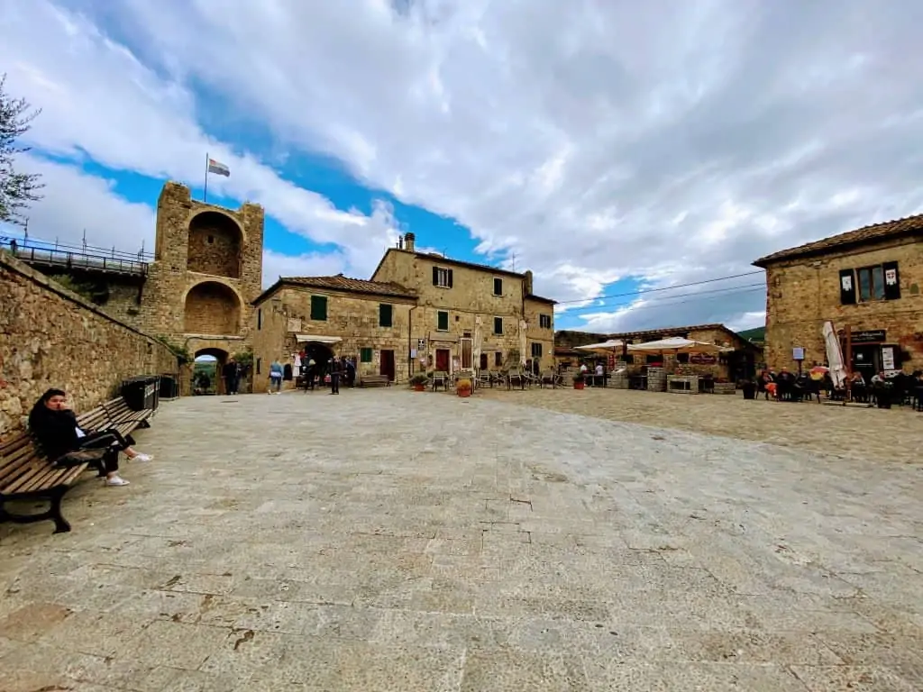 Piazza Roma almost empty on a sunny day with fluffy clouds in Monteriggioni, Italy.  A woman sits on a bench on the left of the piazza.