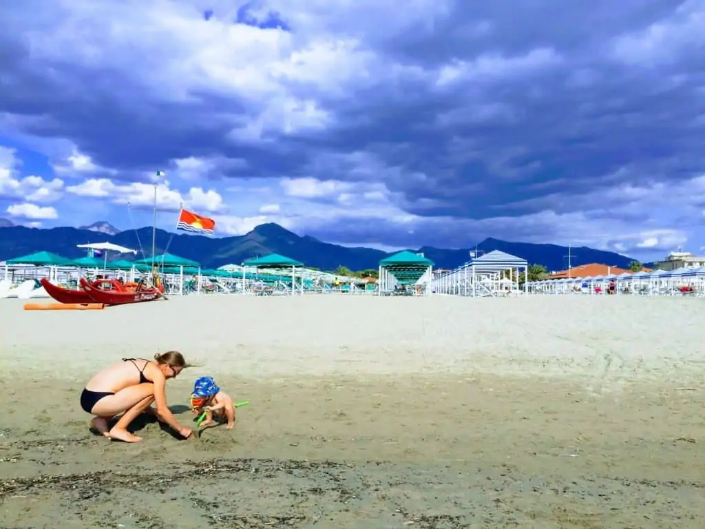 mom and son playing in the sand at beach in forte dei marmi, italy with beach club and mountains in background on a cloudy day
