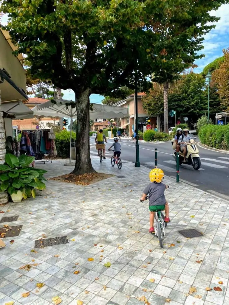 Man and two boys cycling on a wide stone sidewalk in forte dei marmi, italy.  there is a shop on the left selling clothing and a scooter on the street on the right