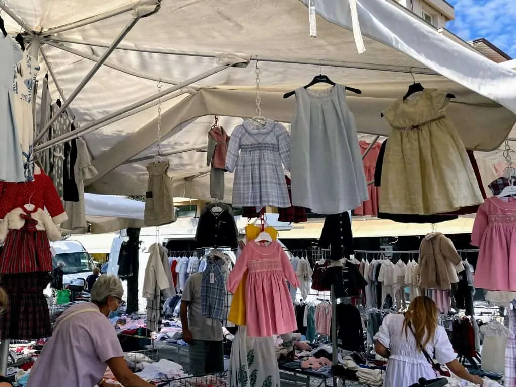 kids clothing stand at the forte dei marmi market.   dresses hang from the top of the tent and there are clothes racks in the background and piles of clothing in the center.  an older woman on the left looks through some of the clothing.