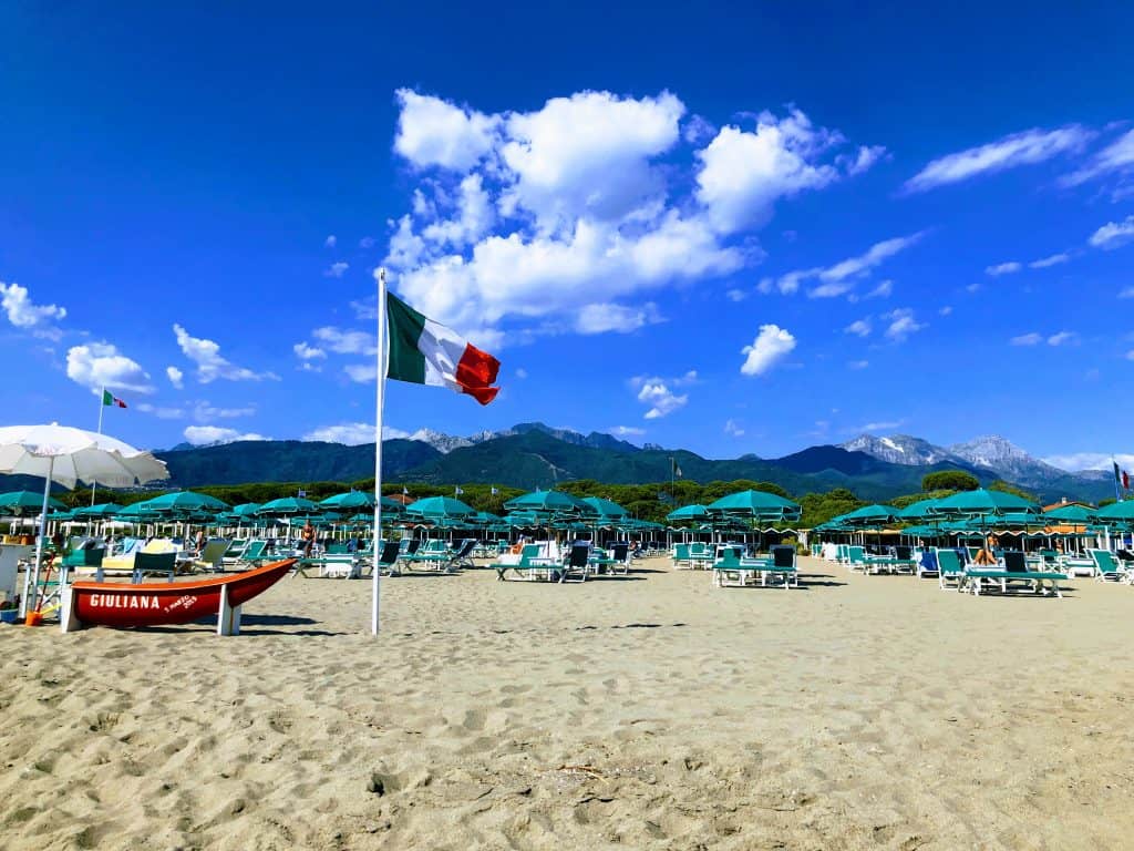 italian flag waving on the beach in forte dei marmi, italy with a red lifesaving boat on the left and beach chairs and umbrellas in the background.  you can see the apuan alps in the distance. it's a sunny day with a few clouds.