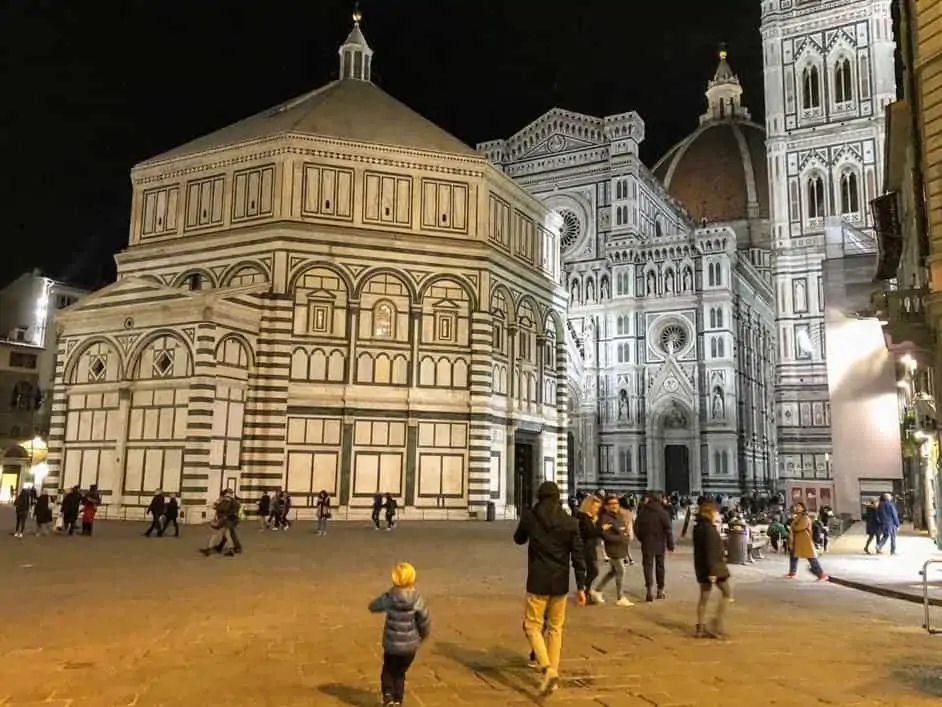 The Baptistry and the Duomo of Florence, Italy at night. They are both lit up and there are a few people walking in the square.