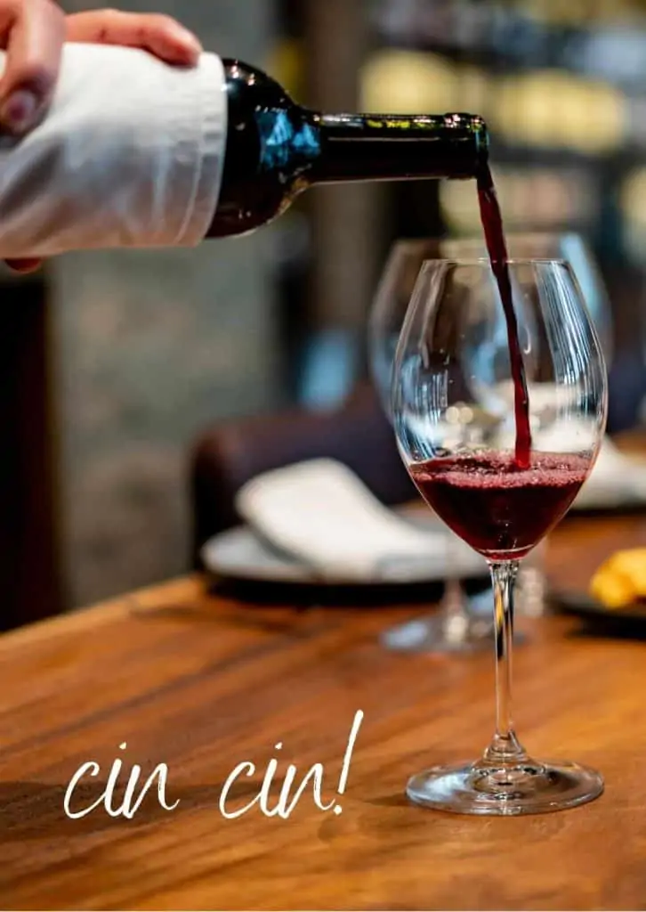 A glass of red wine is being poured from a bottle of wine.  The glass is on a wooden table and you can see a couple of place settings on the table in the background.  The words 'cin cin' are written in white on the bottom left of the photo.