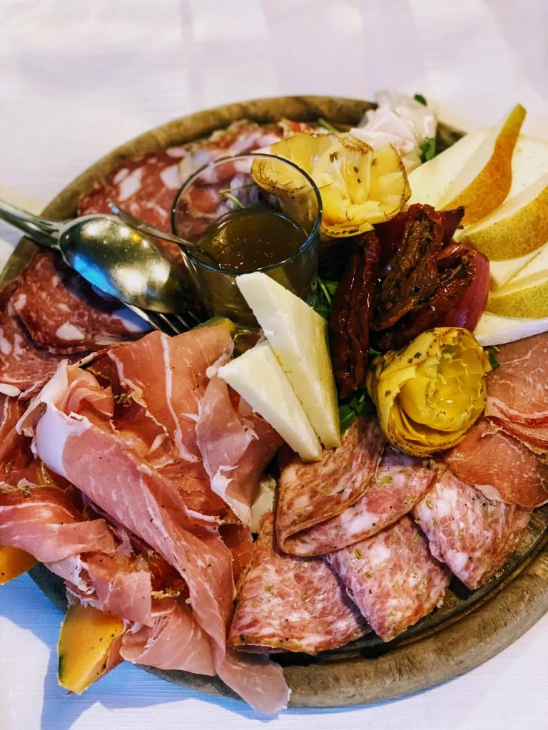 Tuscan antipasto platter on a wooden cutting board on a white table cloth. It contains cured meats, cheese, honey, artichokes, pears, and sundried tomatoes.