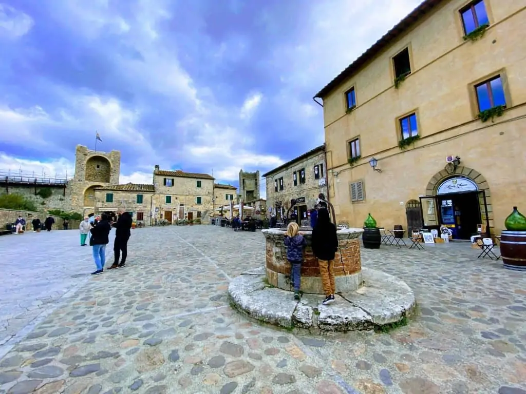 Two boys look into the well in the center of the main piazza in Monteriggioni, Tuscany, Italy.  The sky is blue with puffy white clouds.  You can see the buildings surrounding the square and thre are a few people walking around the square.