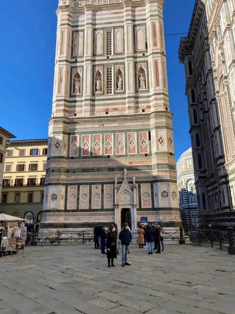 A few people wait in line to enter Giotto's bell tower in Florence, Italy.  You can see lower part of the bell tower in the photo as well as the stone ground of the piazza in the foreground.