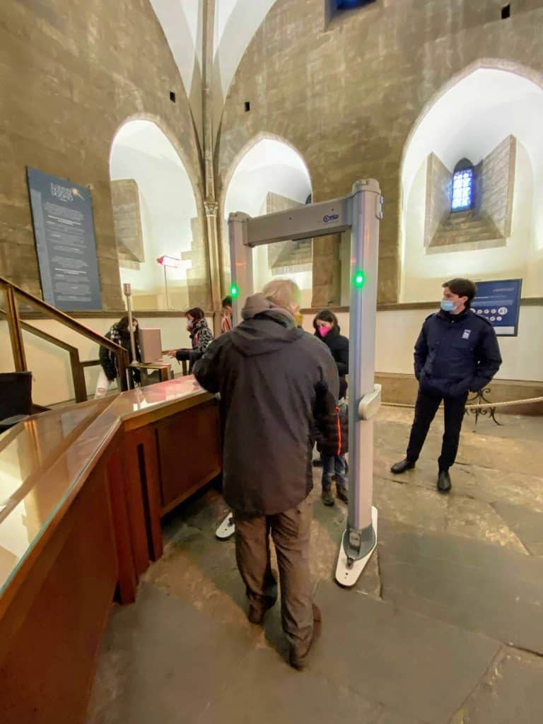 Man is about to walk through a metal detector inside the base of Giotto's bell tower in Florence, Italy. A security guard waits on the other side. Other people have passed through already. Light enters from windows.