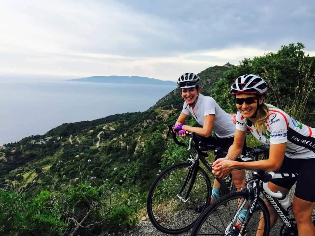 Two female cyclists taking a break from cycling a gravel road on the Monte Argentario peninsula in Tuscany, Italy.  The mountainside is a lush green and there are purple wildflowers.  You can see the sea and another island in the background.
