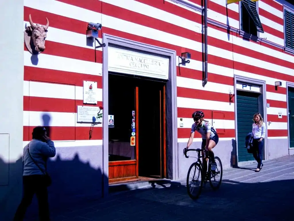 Storefront of Antica Macelleria Cecchini butcher shop in Panzano in Chianti, Italy.  The building has thick red and white stripes on the front and a bull head in the upper left.  There is a cyclist and two pedestrians in front of the shop.
