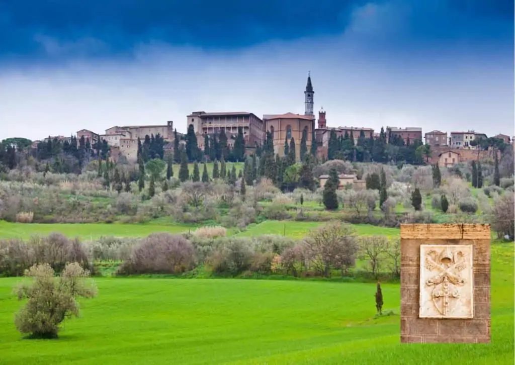 Pienza, in Tuscany, Italy with green fields and woods/brush in the foreground.  In the lower right is the coat of arms of Pope Pius II, who designed Pienza as a utopian city.