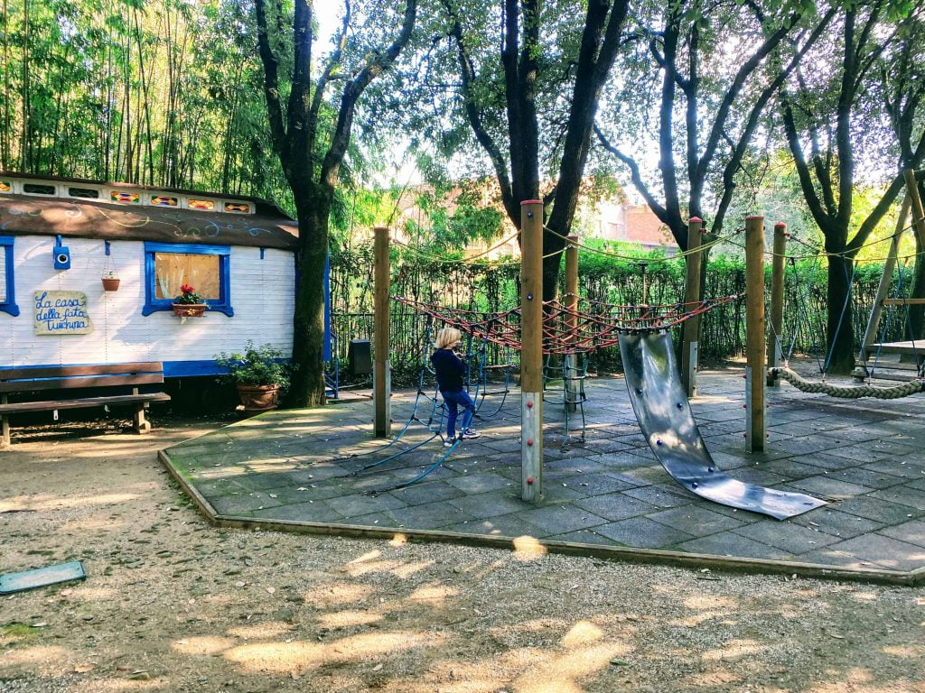 Boy plays on small playground at the Pinocchio Park in Collodi, Italy.  There are rope climbing structures.  There are talle trees shading the area.  On the left is a trailer with the fairy's home inside.