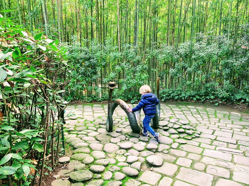 Boy climbs on snake sculpture in Pinocchio Park in Collodi, Italy.  He's in a bamboo forest and the ground is cobblestone.