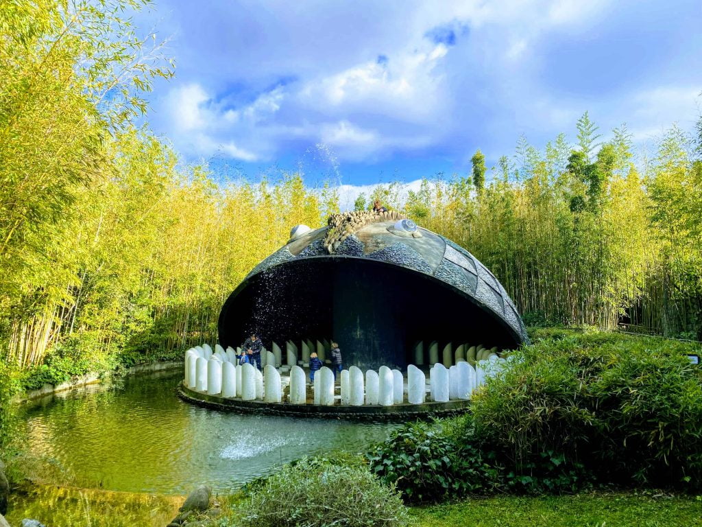 Dogfish sculpture at Pinocchio Park in Collodi, Italy. Surrounded by bamboo forest. You can see people walking inside the mouth near the teeth. The dogfish is spraying water from its spout into the large pond in front of it.