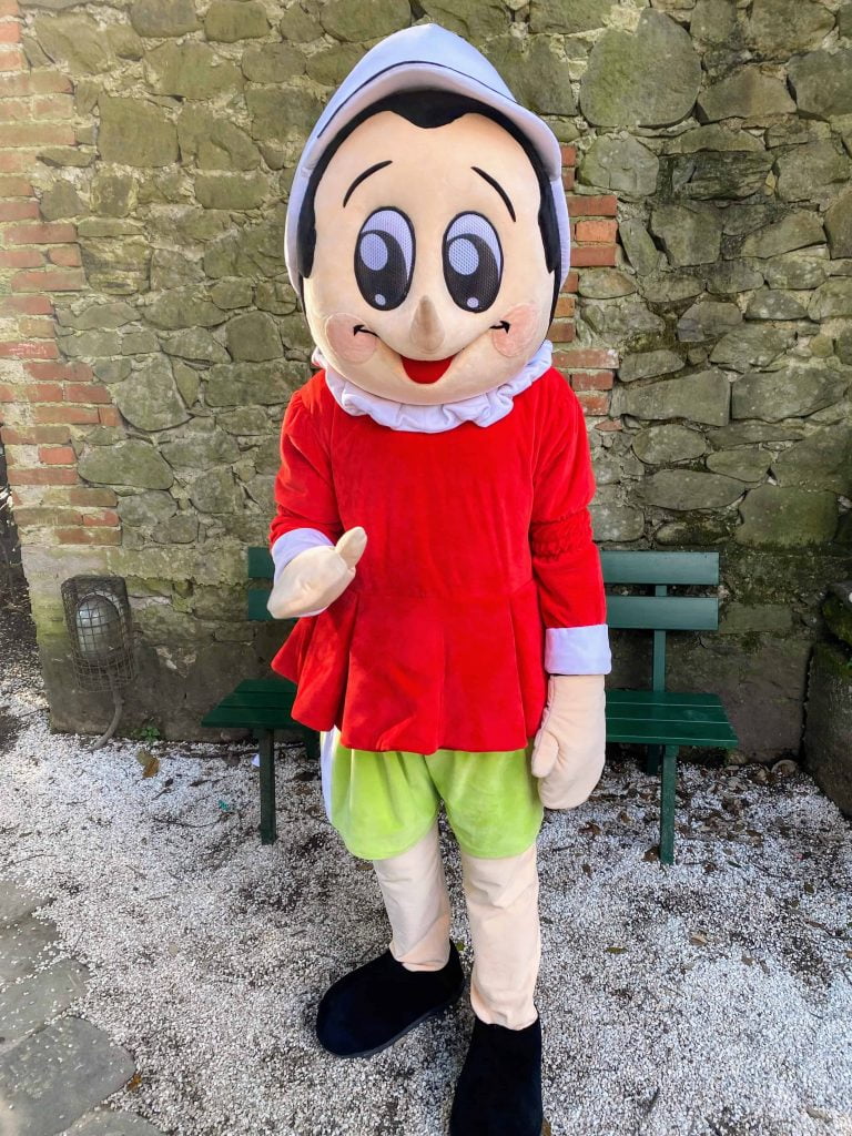 Pinocchio character at the Pinocchio Park in Collodi, Italy.  He's standing alone in the gravel against a stone wall.  There is a green bench behind him.