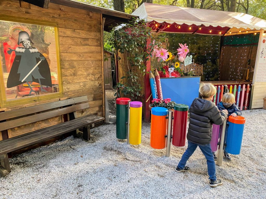 Two boys play a colorful outdoor drum set at the Pinocchio Park in Collodi, Italy.  There are wooden buildings in the background.  One looks like a small stage and the other has a Darth Vader Pinocchio decal in the window.