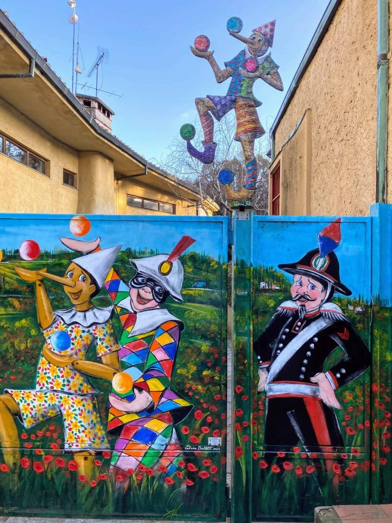 One of many colorful murals in Collodi, Italy, the home of the Pinocchio Park.  The mural is on a service door and shows Pinocchio juggling with a jester while an officer watches.  The background of the mural is the Tuscan countryside with red poppies and a blue sky.  Above the mural is a metal cutout of Pinocchio juggling.  You can see a yellow building in the background of the photo.