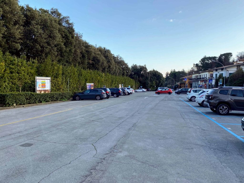 Large parking lot, about half full, at the Pinocchio Park in Collodi, Italy.  There are tall trees and bushes bordering it to the left and two-story buildings on the right.
