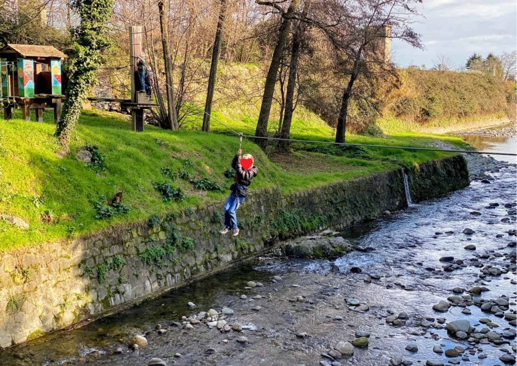 Boy ziplines across small river at the Pinocchio Park in Collodi, Italy.  The hillside on the left has green grass and platforms of the adventure park.  The river is shallow with stones showing above the water.  There are a couple of trees in the grass on the far bank.