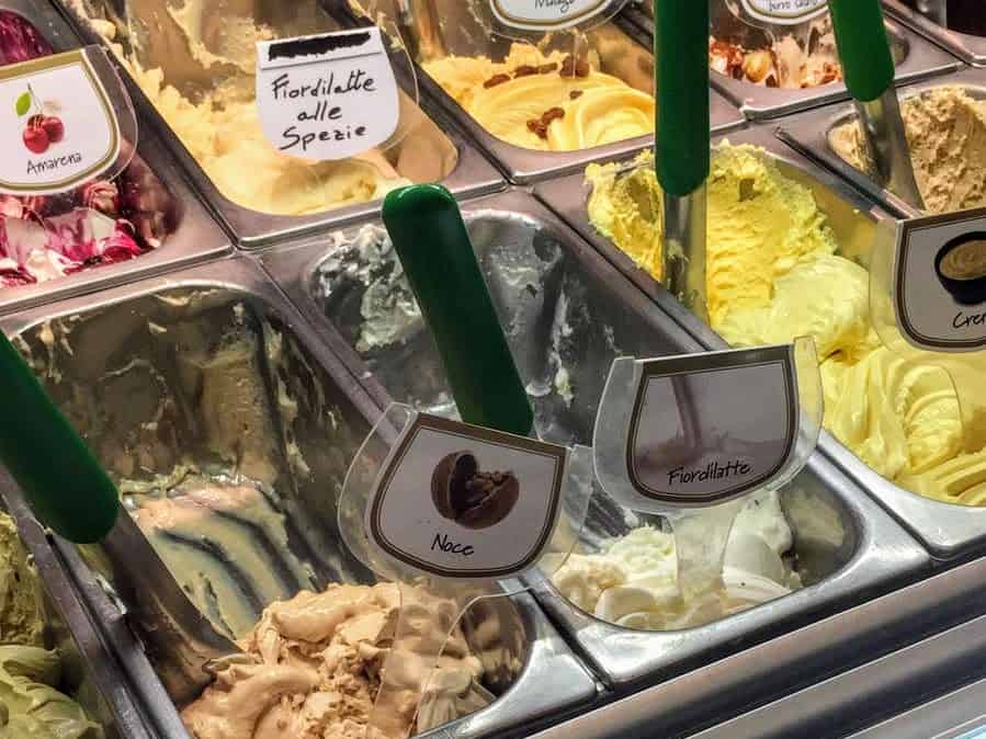Close up of gelato in containers in a shop.  You can see amarena, fiordilatte alle spezie, noce, fiordilatte, and crema.  There are also the serving paddles in a few containers and the paddles have green handles. 