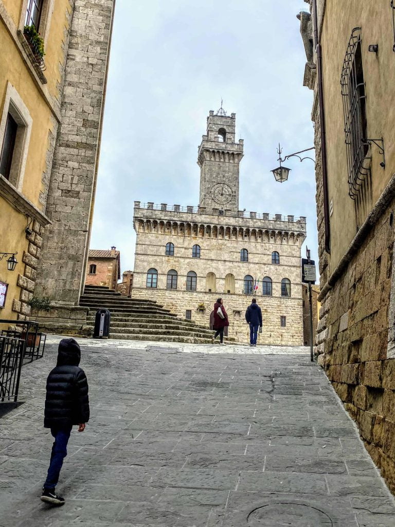 Boy walking toward the clock tower of the Palazzo Communale in Montepulciano Italy.  There are only two people in the piazza ahead.