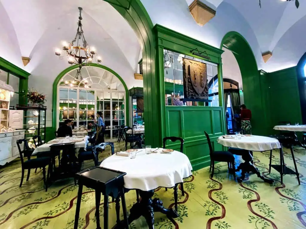 Dining room at Gucci Osteria in Florence, Italy.  Yellow floors with painted leaves and ribbons.  Green walls and mirrors.  Round tables with white tablecloths and black chairs.  There is one couple dining.