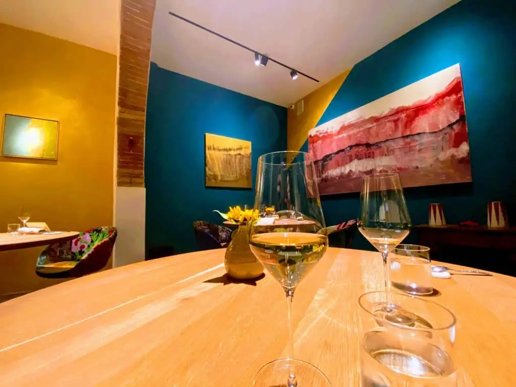 Table at Linfa Restaurant in San Gimignano, Italy. It's one of Tuscany's Michelin star restaurants. You can see two glasses of white wine on the wooden table.  The walls are painted blue and yellow and there is colorful art on the walls.  There is no one in the dining room.