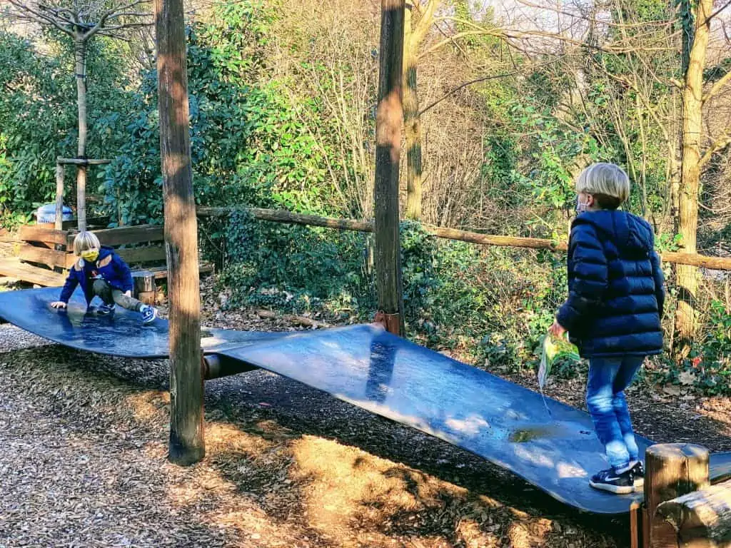 Two boys playing on a playground toy in a forested area of the Pistoia Zoo (Zoo di Pistoia).  It's a large black mat sitting off the ground with the help of logs and wooden poles.  You jump on one side and it makes the other side go up.