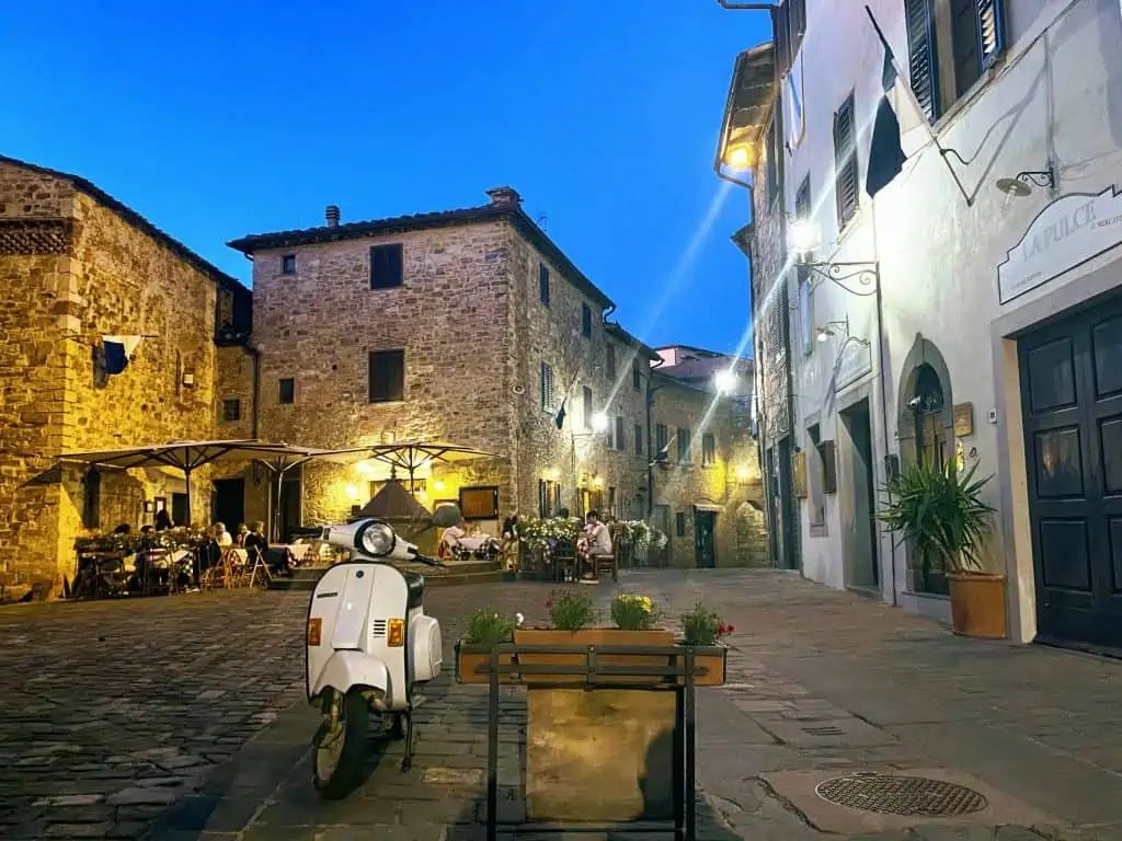 Piazza at night in Italian village. You can see a vespa and people dining outdoors.