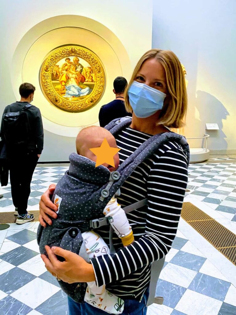 Mom holding child in baby carrier at Uffizi Gallery in Florence.  You can see a painting by Michelangelo in background.