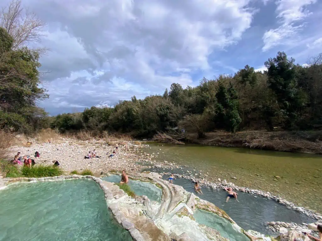 View of the Bagni di Petriolo hot springs in Southern Tuscany, Italy.  There are a few people soaking and you can see the river and trees in the background.