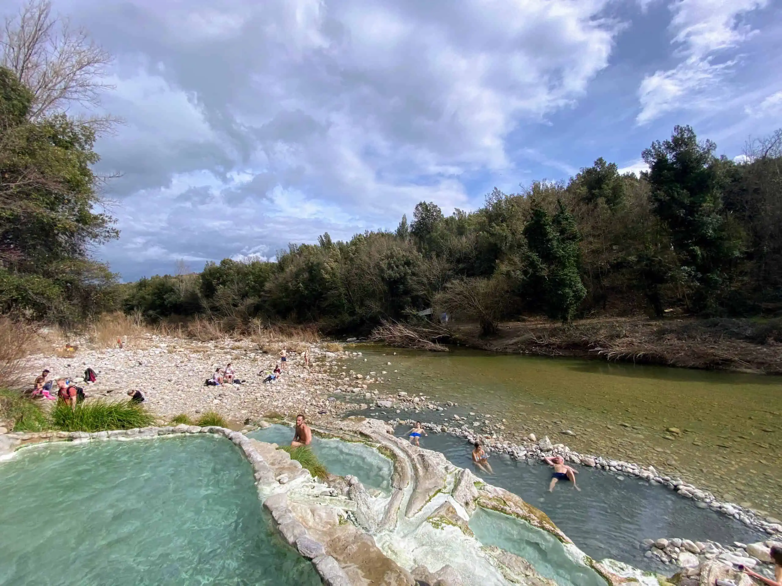 Turquoise hot springs with a river and forest in the background. Sunny day with fluffy white clouds. People soaking in hot springs.
