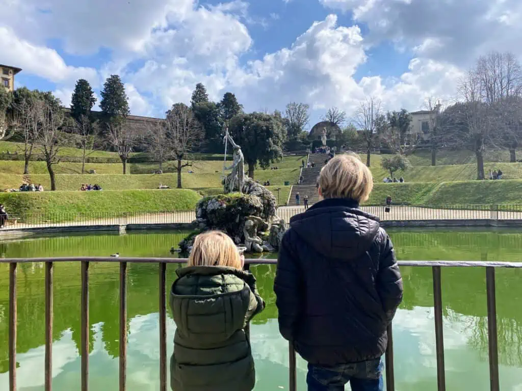 Boys looking out at fountain with statue of Neptune in the middle.  Terraced grassy hills in background and a few trees.  Sunny day with puffy white clouds.