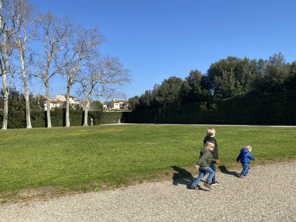 Boys running on gravel path in Boboli Gardens in Florence, Italy.  You can see large grassy area and tall trees in the background, plus a few apartment buildings in the distance.