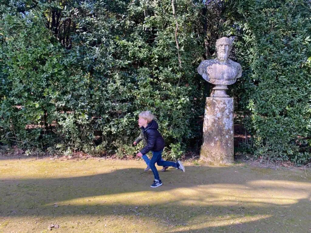 Boys running on dirt path past statue.  Tall bushes behind the statue.