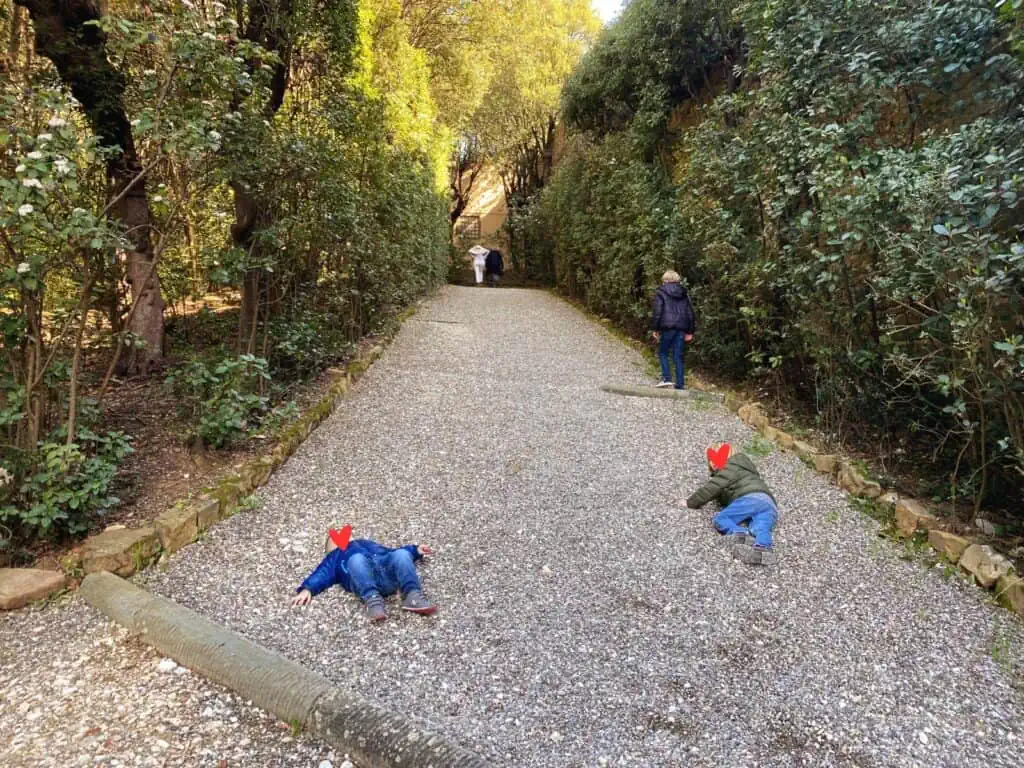 Boys laying on gravel path in Boboli Gardens.  The path is lined with bushes and trees.  There is another boy on the path ahead of them.