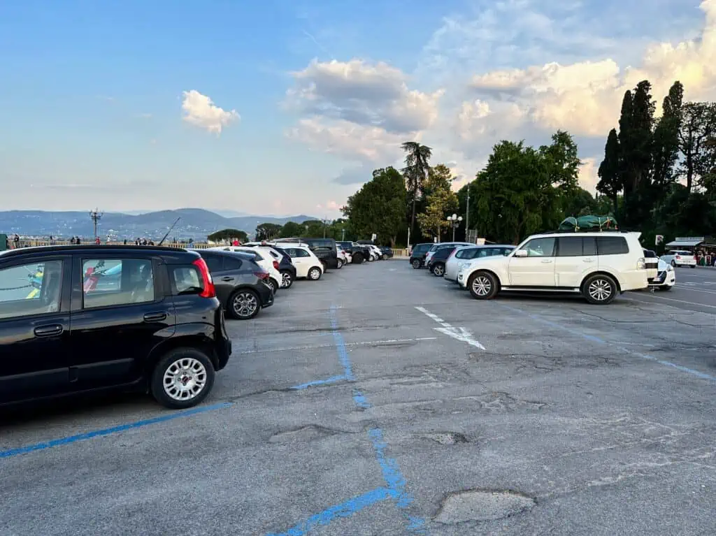 Parking area in Piazzale Michelangelo in Florence, Italy. It's full of cars. You can see tall trees in the background.
