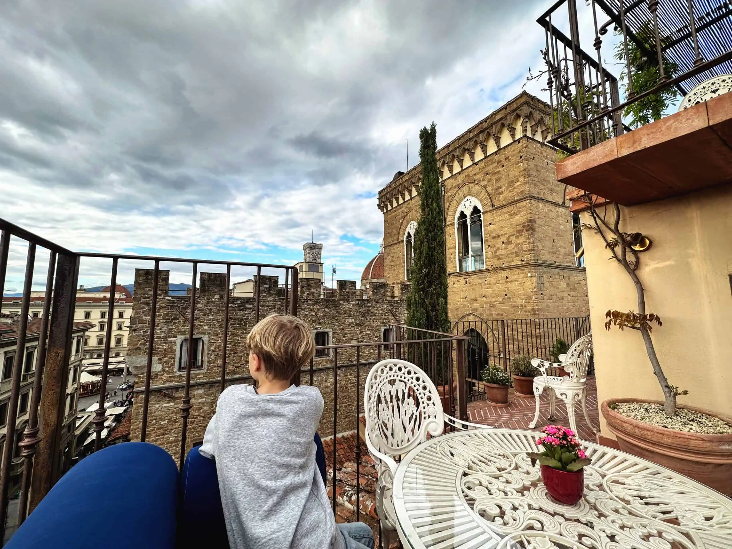 Boy in grey sweatshirt looking at view of top of buildings in Florence, Italy. He's sitting on a blue chair at a white table. You can see part of the Duomo dome and Piazza della Repubblica.