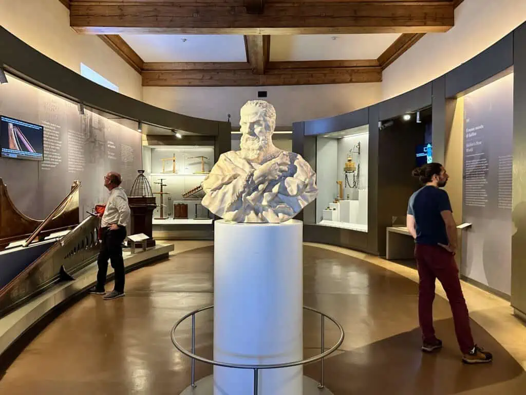 Bust of Galileo in the center of a room his namesake museum in Florence, Italy.  Two men are looking at exhibits in the room.