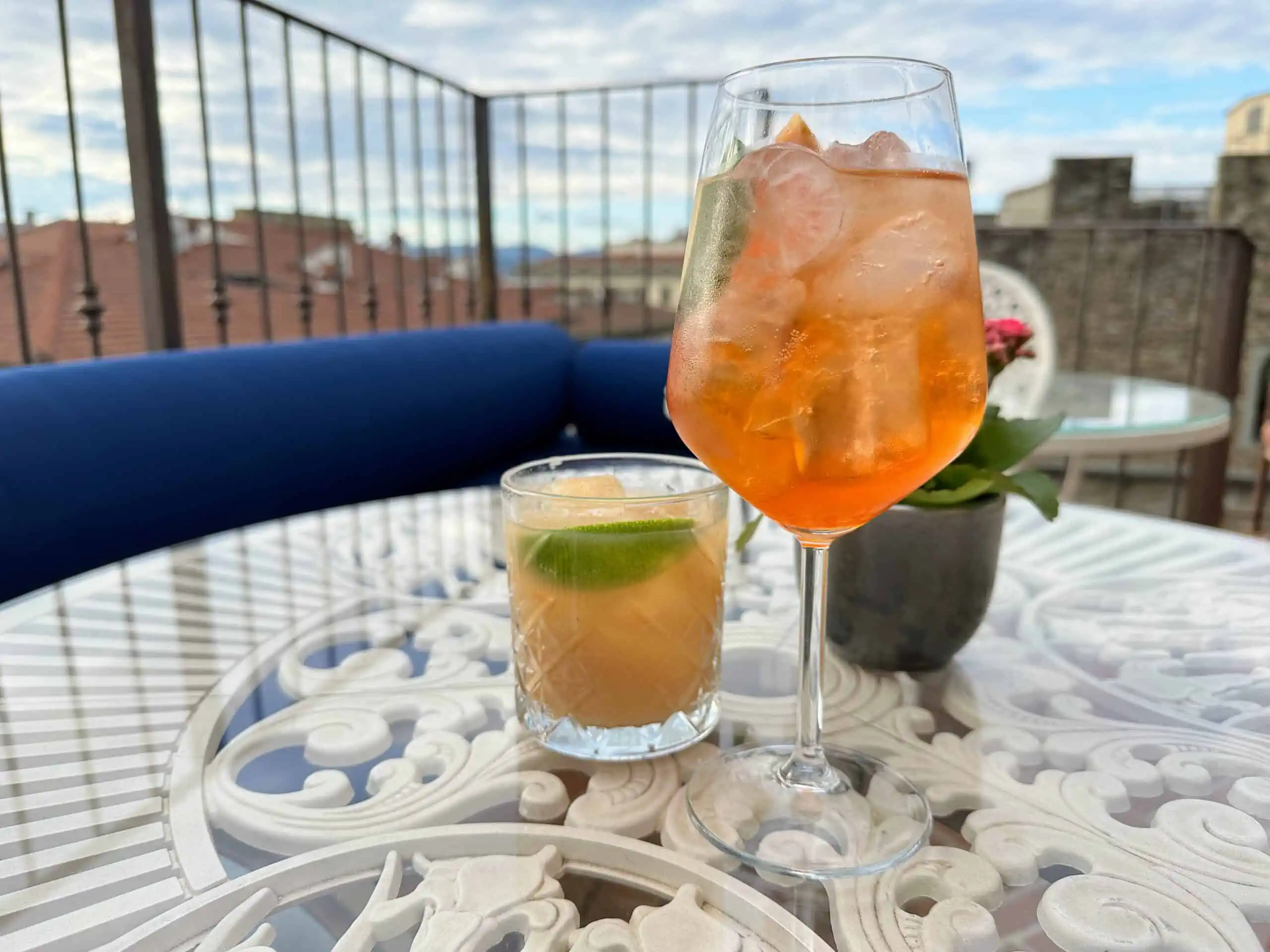 Close up of spritz and juice on a white table at a rooftop bar. You can see the metal railing and city buildings of Florence in background.