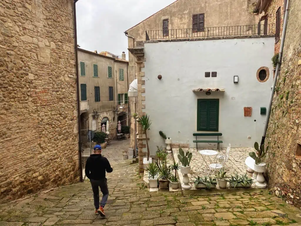 Man walking down a cobblestone hill. Buildings on both sides and ahead. It's a small village in Tuscany. Terrace with table and chairs on the right with plants and cacti.