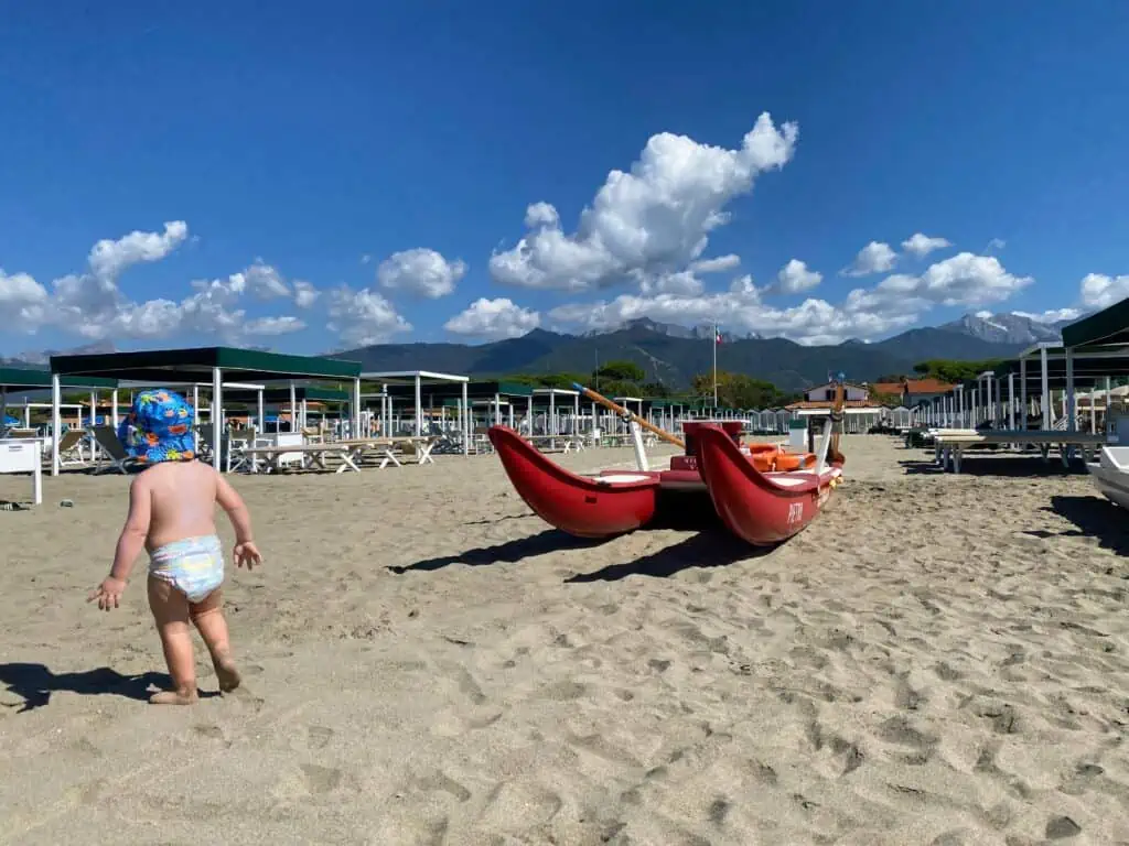 Boy walking away from camera at beach in Italy. You can see mountains in the background and a red lifeguard boat in the photo. There are beach tents and lounge chairs set up.