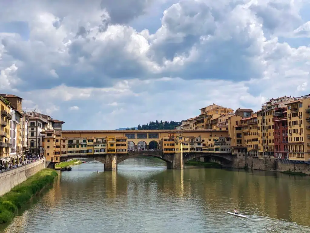 View of Florence, Italy's Ponte Vecchio bridge on a sunny day. You can see puffy white clouds and there are buildings on both sides of the river.