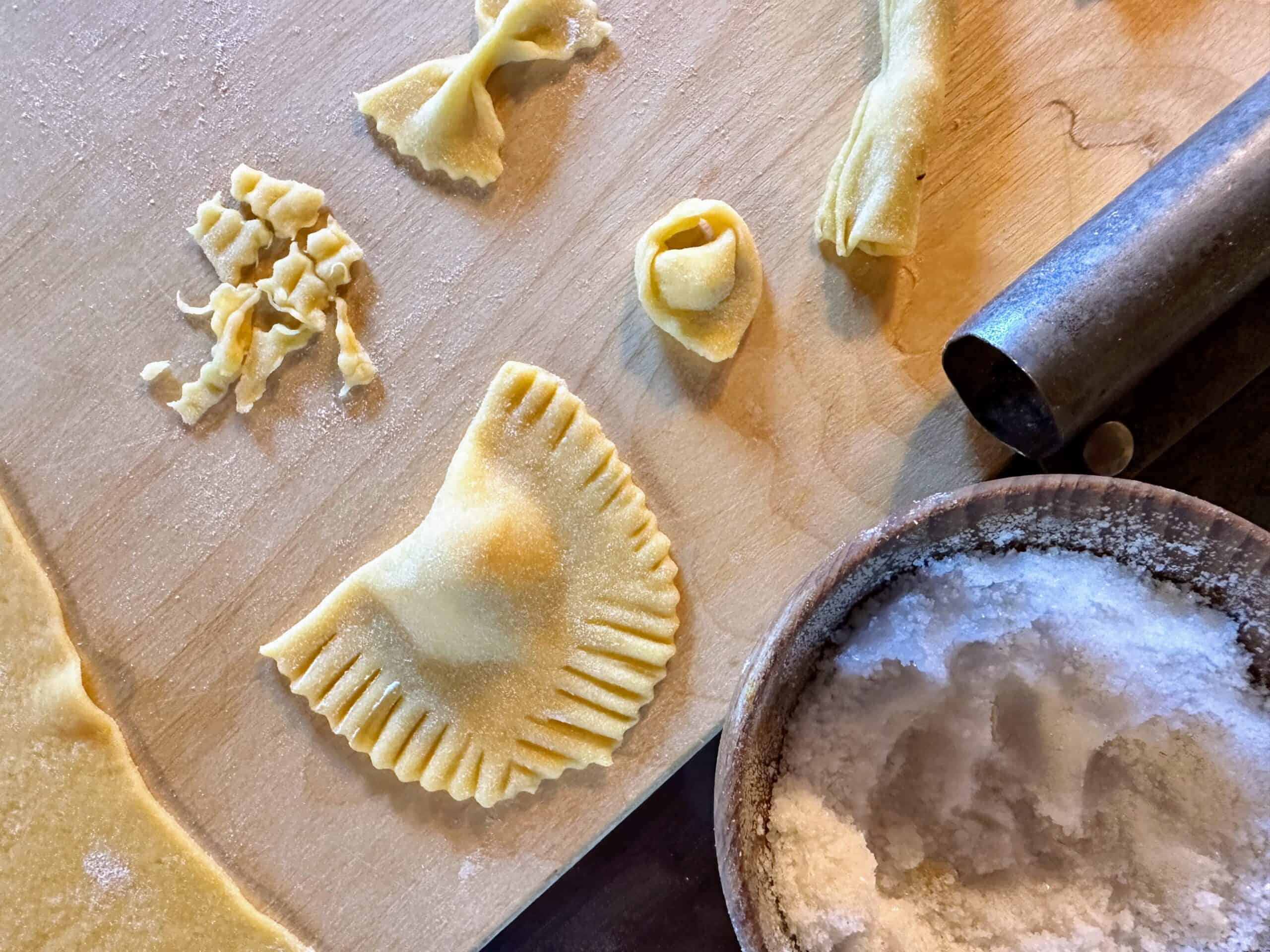View of different fresh pasta shapes on wooden cutting board. Also small container of salt in lower right.