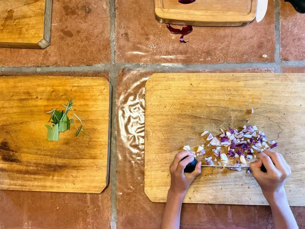 Kid uses mezzaluna to chop onion on a wooden cutting board.