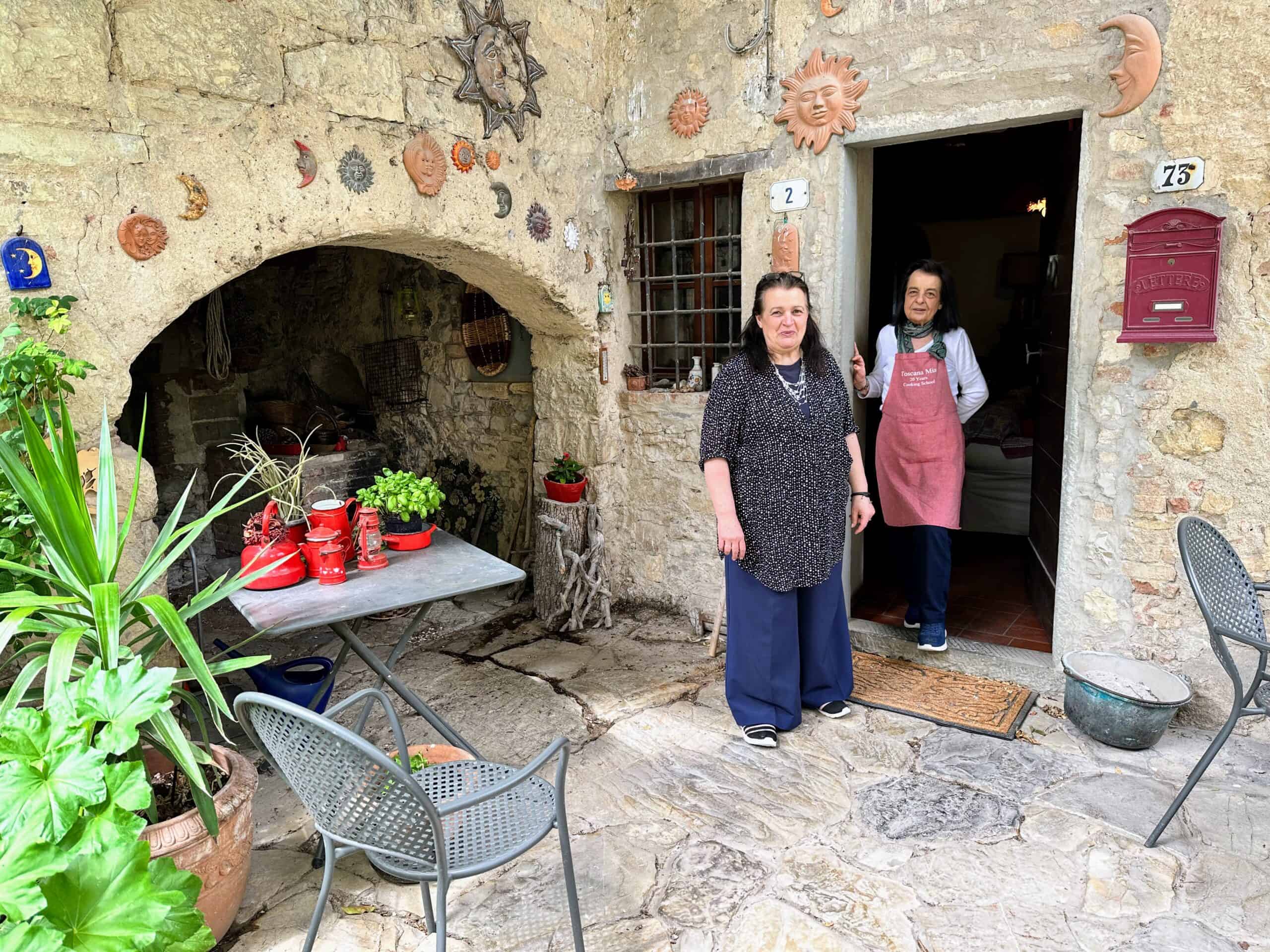 Two women stand in front of entrance to home. Building is made of stone, there are decorations on the walls outside. Plants and chairs and table next to them on front terrace.