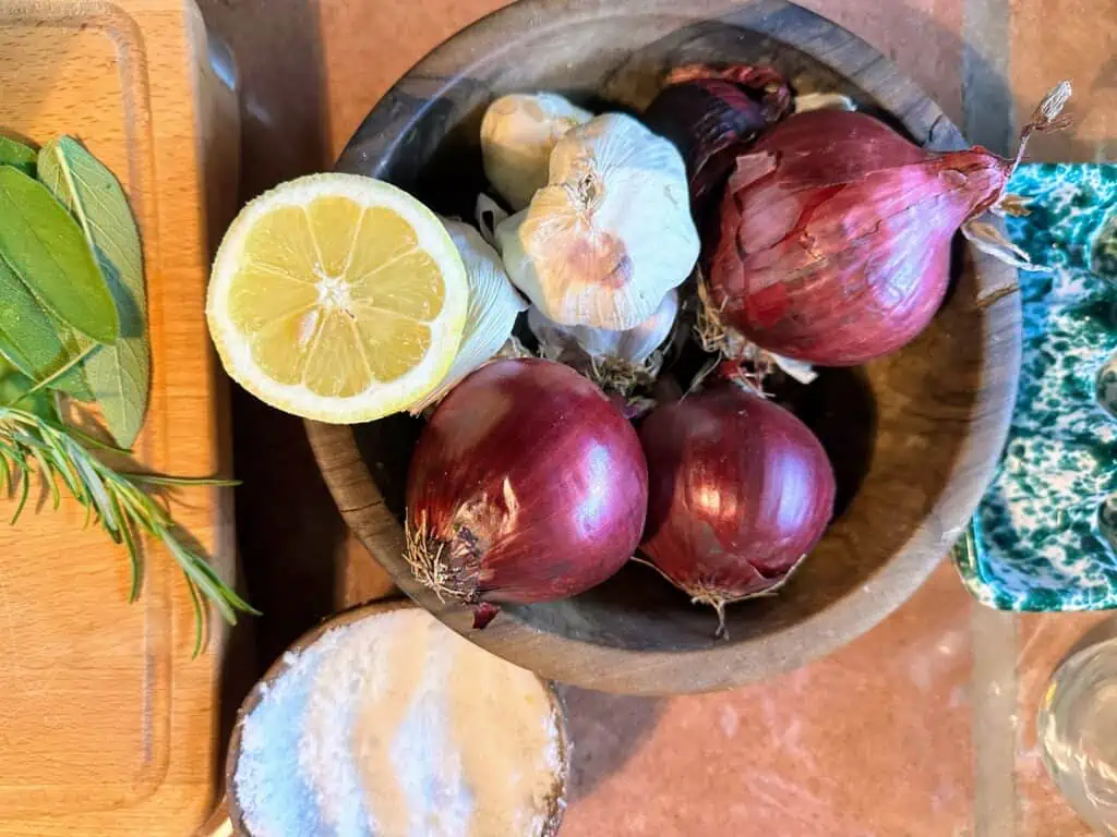 Wooden bowl with lemon, garlic, and red onions. Surrounding it are fresh sage and rosemary and a dish of salt.