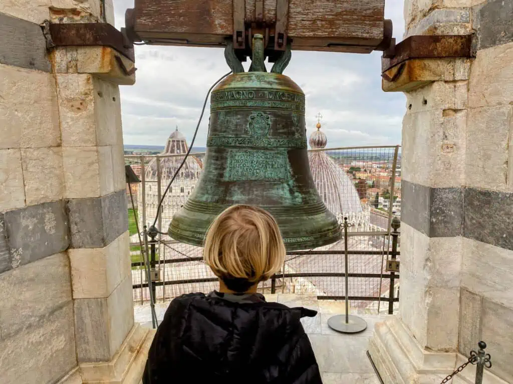 Boy looking at bell at the top of the Leaning Tower of Pisa. You can see baptistery and Duomo in the background.