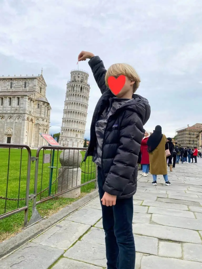 Boy in black coat and jeans posing with the Leaning Tower of Pisa. There are people in the background.