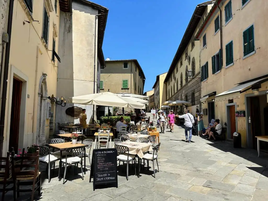 Pedestrian street in Castellina in Chianti. People are walking and also sitting outdoors under umbrellas at a restaurant.