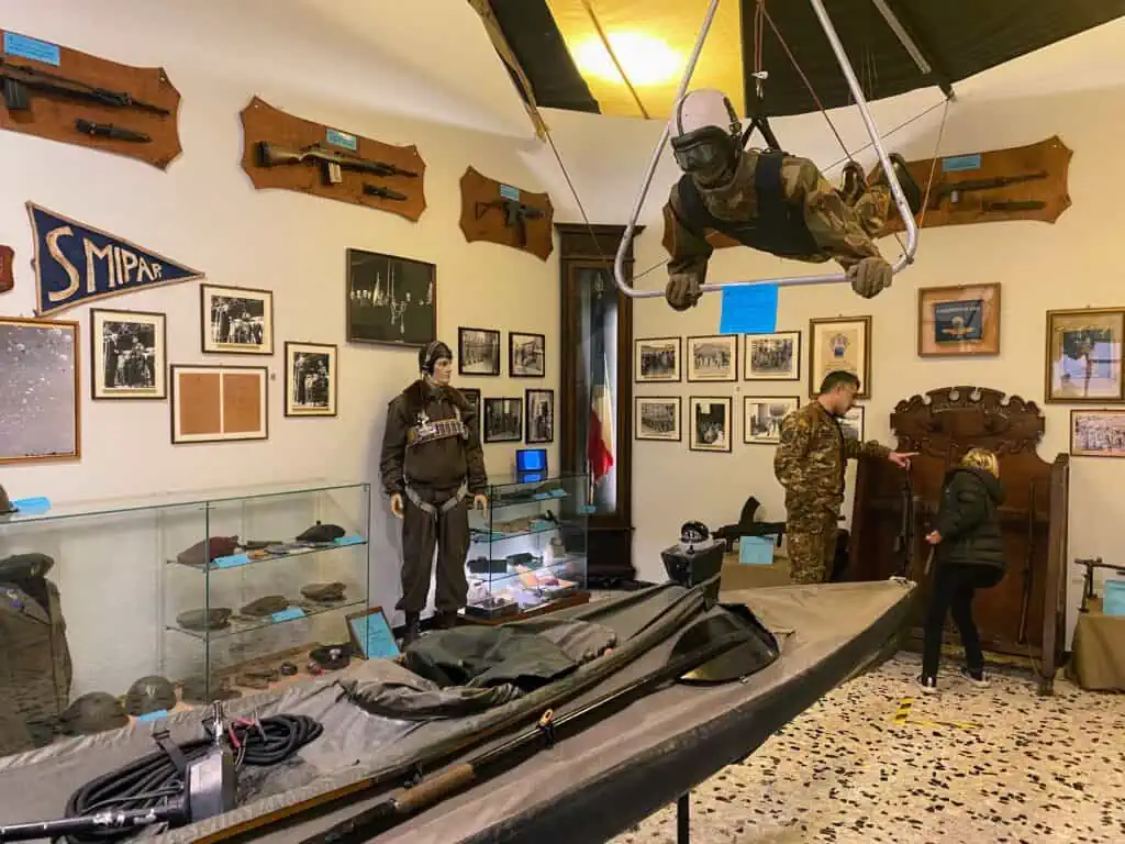 Boy speaks with paratrooper inside Italian Army paratrooper museum in Pisa, Italy. You can see weapons, photos, uniforms, a kayak, helmets, and more artifacts in the room.